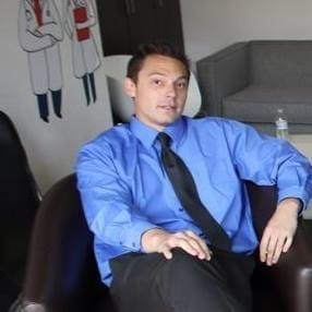 A man in a blue dress shirt and black tie sits on a dark chair with one leg crossed over the other. Behind him are a gray sofa, a table with a water bottle, and a wall with cartoon drawings of people in lab coats.
