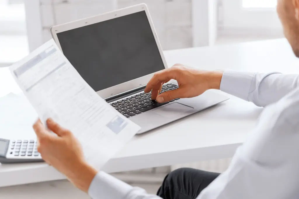 A person sitting at a desk uses a laptop while holding a document, with a calculator nearby. The person's face is not visible, focusing attention on their hands and work materials.