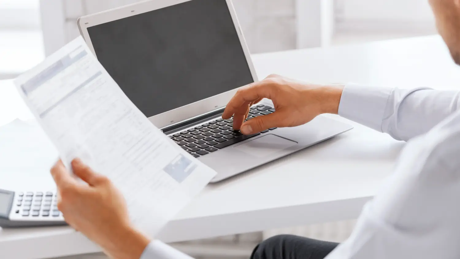 A person sitting at a desk uses a laptop while holding a document, with a calculator nearby. The person's face is not visible, focusing attention on their hands and work materials.