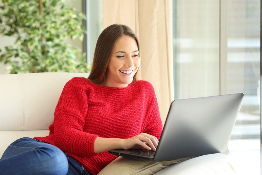 Woman in a red sweater sitting on a couch, smiling and typing on a laptop, with a window and green plant in the background.