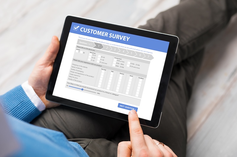 A person in a blue sweater is holding a tablet displaying a brand reputation management survey screen, with their finger on the "next question" button.