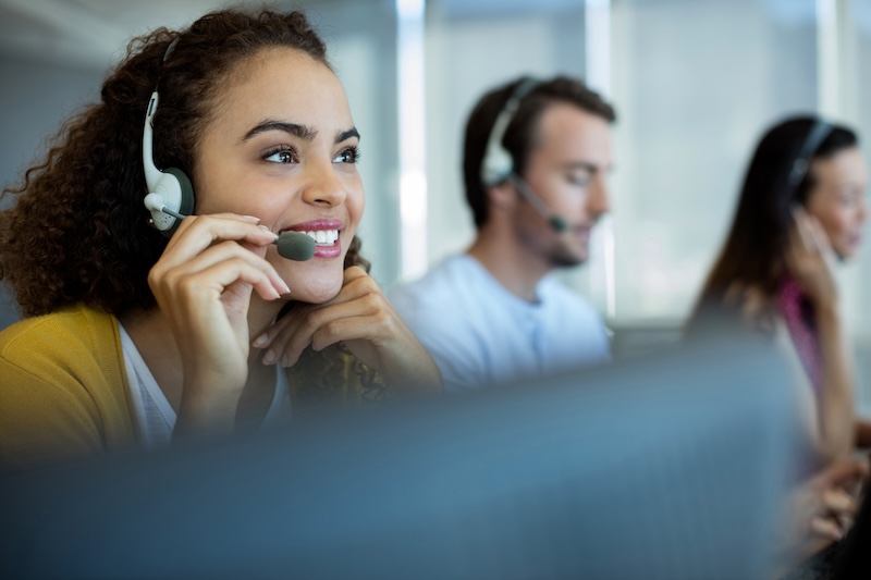 A young woman wearing a headset and smiling while working in a bustling call center focused on brand reputation management, with colleagues also equipped with headsets in the background.