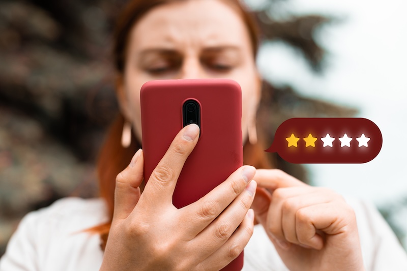 A woman with red hair, holding a smartphone and touching the screen, with an animated graphic showing a one-star rating floating beside the phone, highlighting the challenges of brand reputation management.