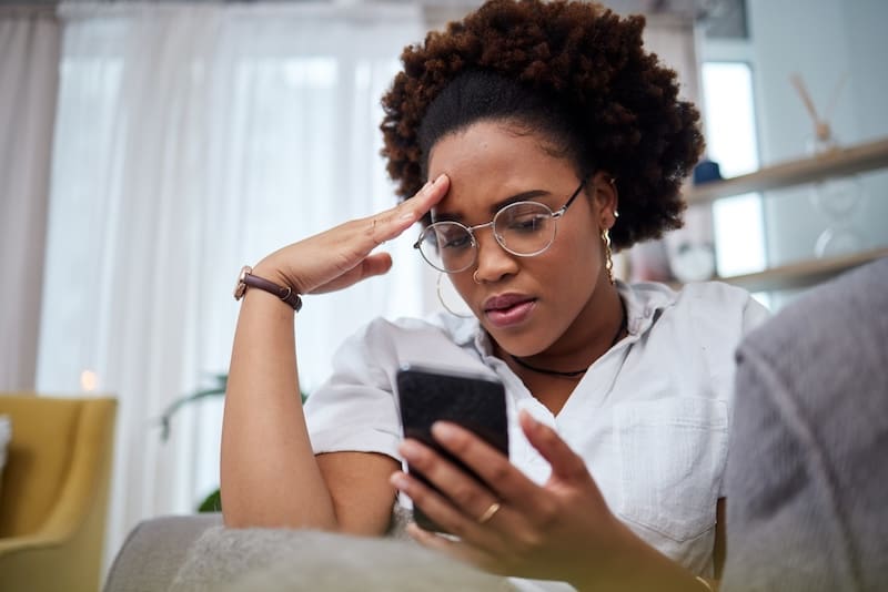 A young woman with glasses and a curly updo looks frustrated or confused while staring intently at her smartphone, possibly managing brand reputation issues, in a brightly lit living room.