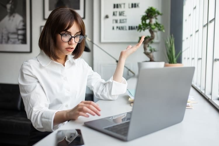 A woman in a white shirt and glasses gesturing with her hand while looking at her laptop screen with a puzzled or concerned expression over unwanted Google search results in a modern office setting.