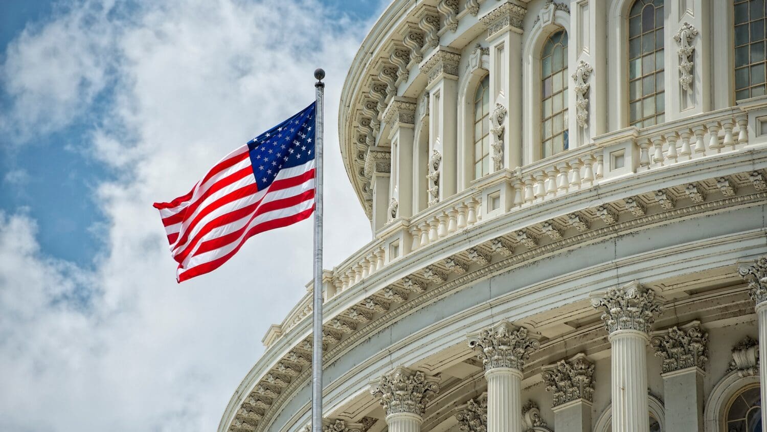 The U.S. flag waves in front of the ornate white columns and windows of the United States Capitol building, where leaders discuss issues like harmful online content, under a partly cloudy sky.