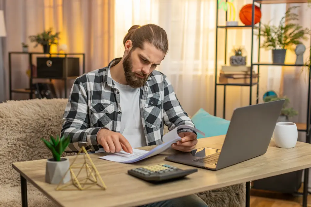 A man with a beard sits at a desk at home, looking at papers. A laptop, calculator, mug, and plant are on the desk. Shelves, plants, and soft lighting are in the background.