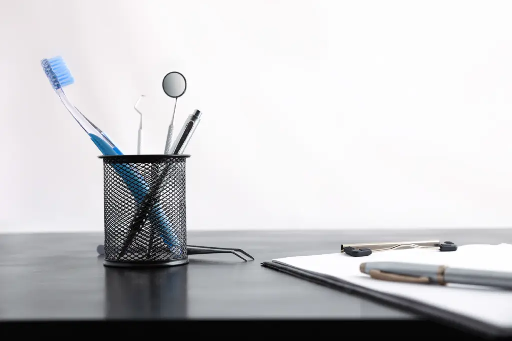 A metal mesh pen holder with toothbrush, dental mirror, and dental tools sits on a black desk next to a clipboard with papers and a pen, against a plain white background.