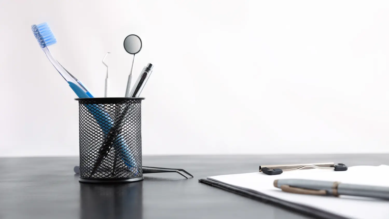 A metal mesh pen holder with toothbrush, dental mirror, and dental tools sits on a black desk next to a clipboard with papers and a pen, against a plain white background.
