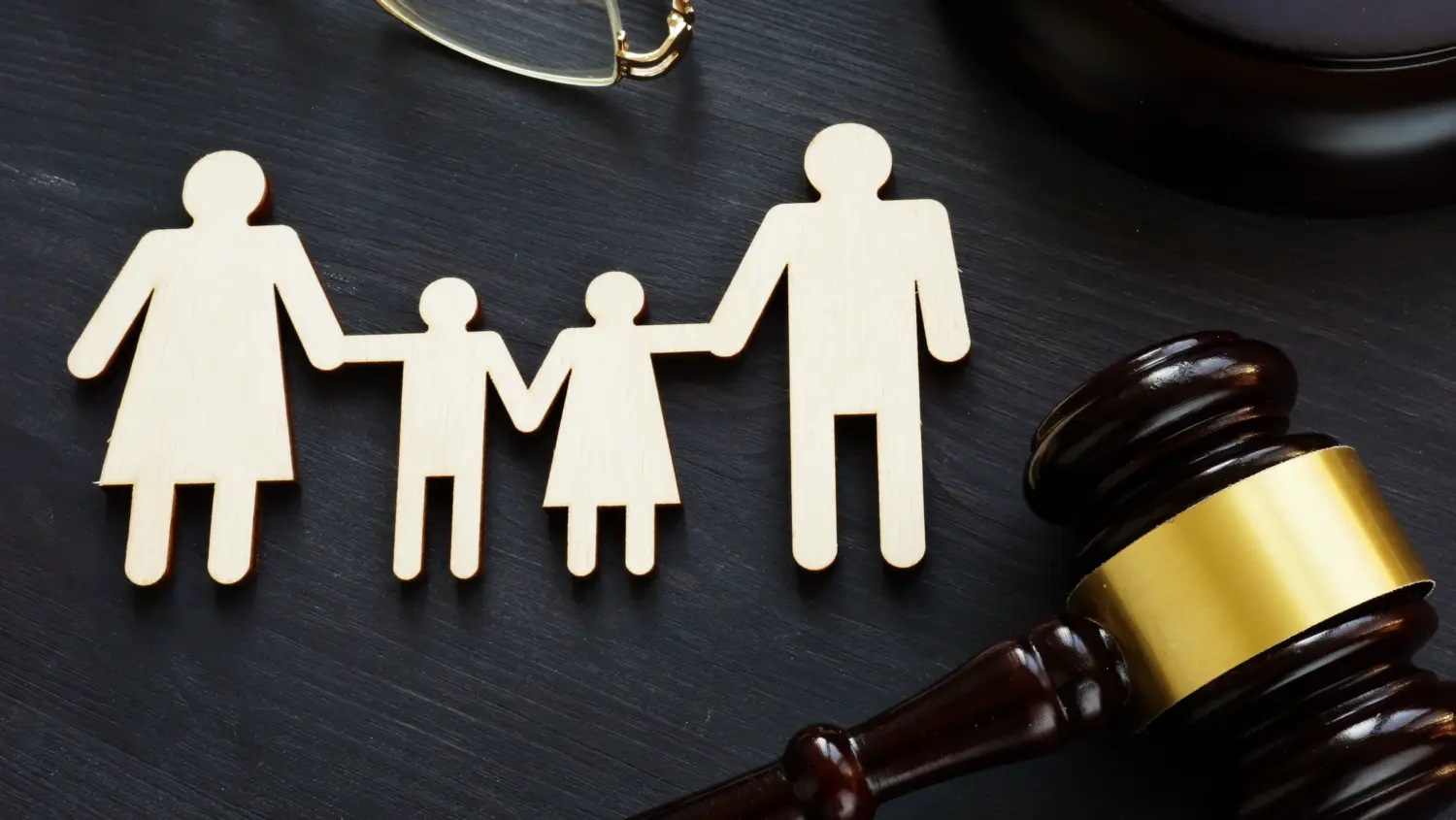 Wooden cutouts of two adults and two children holding hands are placed on a dark surface near a judge's gavel and eyeglasses, symbolizing family law, custody, or legal family matters.