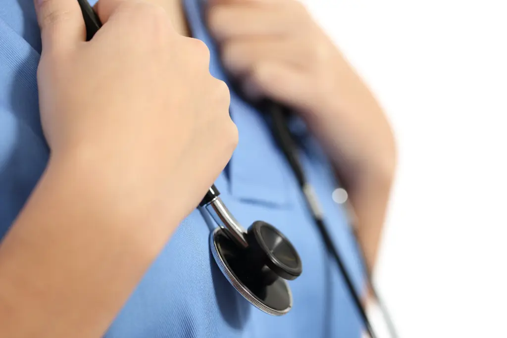 A person in blue scrubs holds a stethoscope against their chest, suggesting a healthcare professional. The background is white and out of focus.