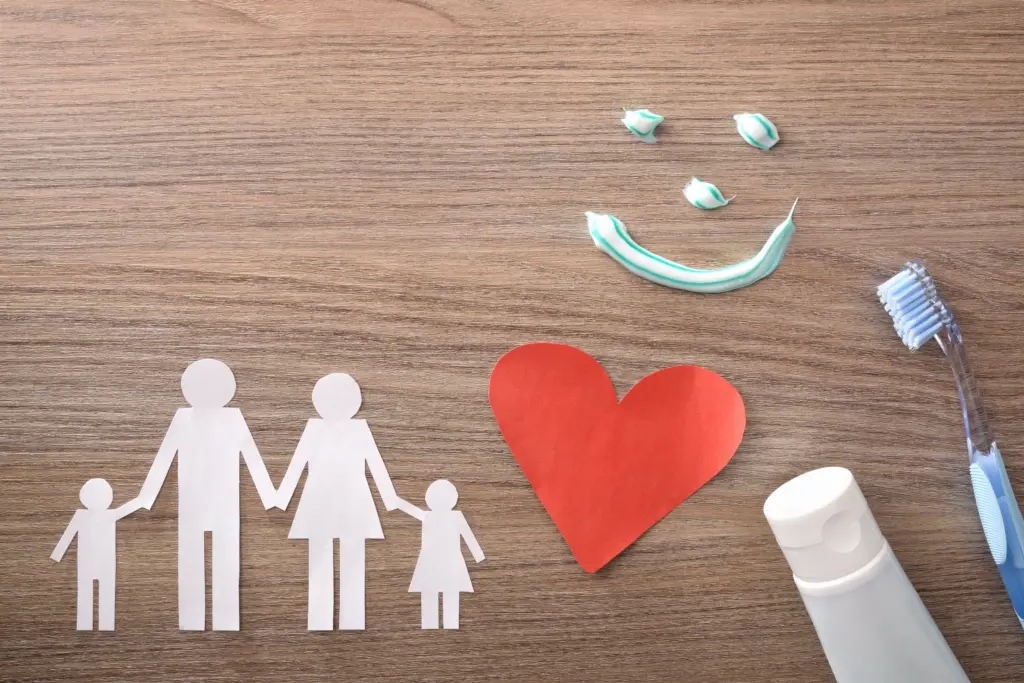 A paper cutout family, red heart, toothpaste tube, toothbrush, and a smiley face made of toothpaste on a wooden surface.