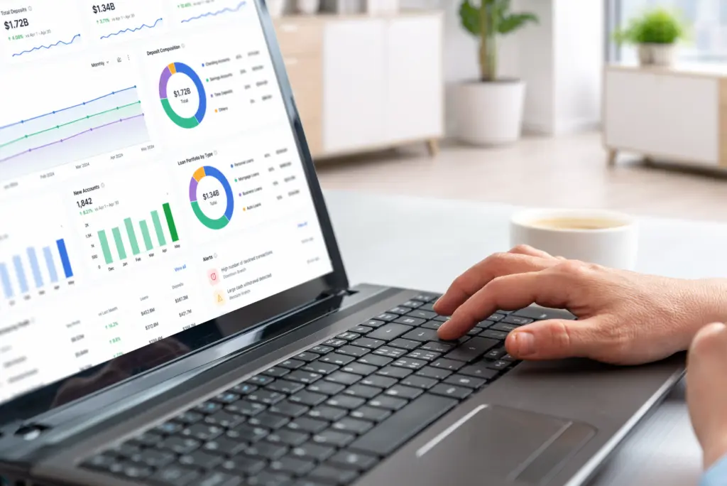 A person uses a laptop displaying colorful financial dashboards with charts, graphs, and analytics in a bright room, with a cup and plant in the background.
