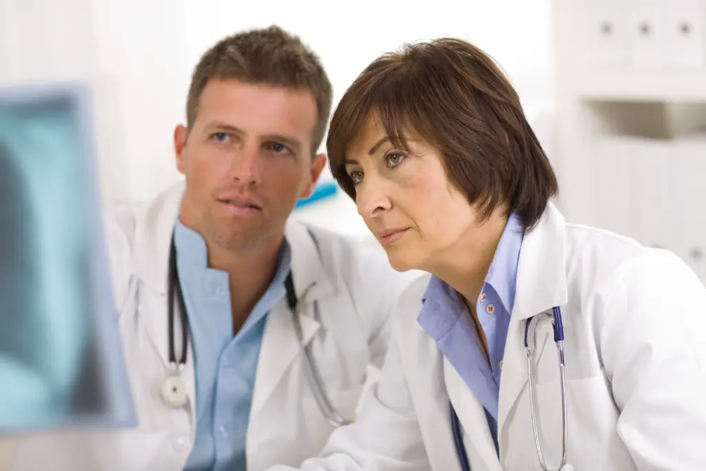 Two doctors in white coats with stethoscopes closely examine something on a computer screen, appearing focused and engaged in a medical discussion. The background is a bright, clinical setting.