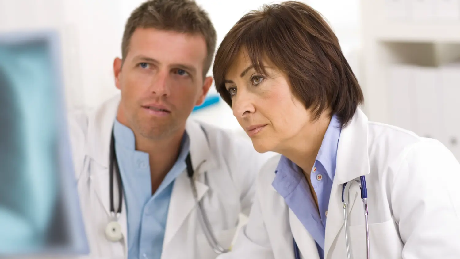 Two doctors in white coats with stethoscopes closely examine something on a computer screen, appearing focused and engaged in a medical discussion. The background is a bright, clinical setting.