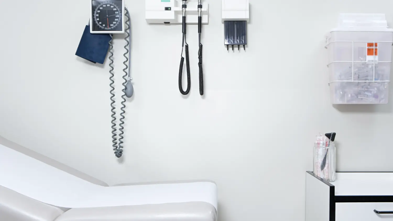An empty medical exam room with a white exam table, blood pressure monitor, otoscope, ophthalmoscope, and other medical tools mounted on the wall, plus a wall organizer with supplies.