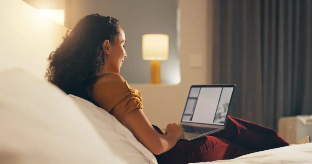 Woman with curly hair sits on a bed, using a laptop and smiling. A lit lamp and blurred background suggest a cozy, relaxed indoor setting.