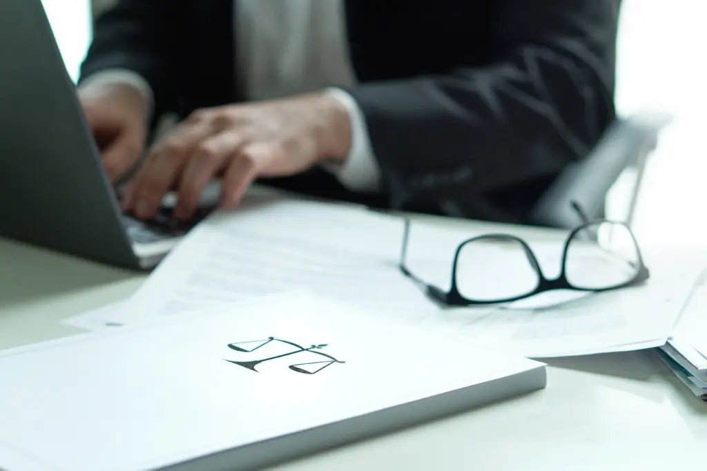 A person in a suit types on a laptop at a desk with documents, eyeglasses, and a folder featuring a scales of justice symbol, suggesting a legal or business setting.