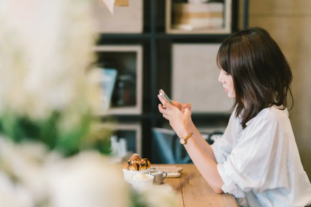A woman with shoulder-length dark hair sits at a wooden table in a café, looking at her smartphone. There is a dessert and a small cup on the table in front of her, with shelves and blurred decor in the background.