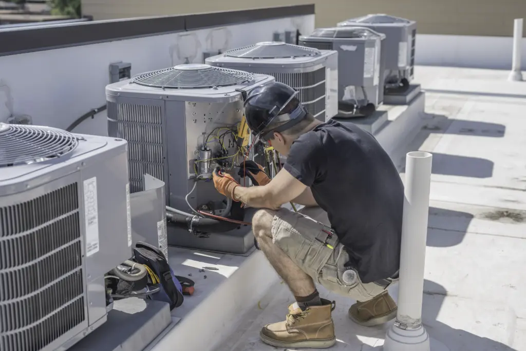 A technician wearing a hard hat and gloves repairs an air conditioning unit on a rooftop, surrounded by several other HVAC units.