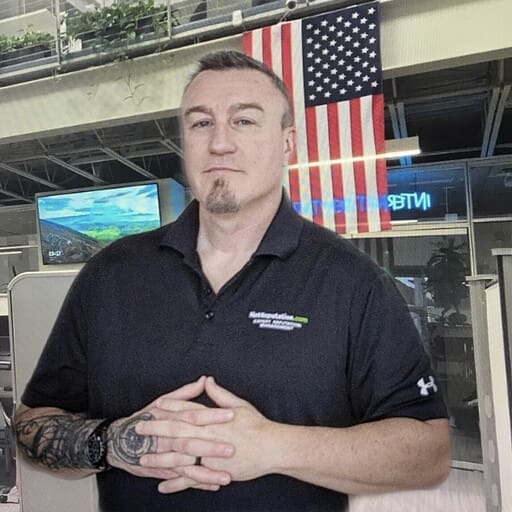 A man with short hair, a goatee, and tattoos on his left arm stands indoors with his hands clasped. He wears a black polo shirt, while an American flag and office furnishings provide a backdrop, suggesting a strong sense of team spirit.