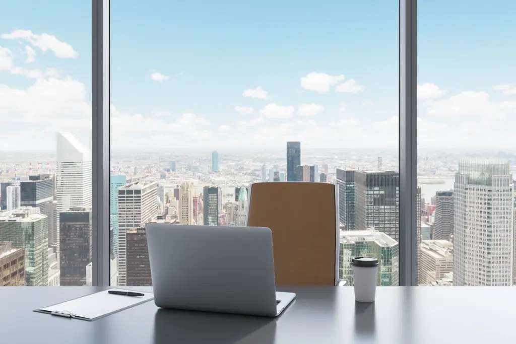 A modern office desk with a laptop, pen, paper, and coffee cup in front of a large window, overlooking a city skyline with tall skyscrapers under a blue sky with scattered clouds.