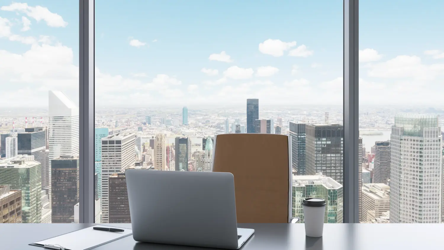 A modern office desk with a laptop, pen, paper, and coffee cup in front of a large window, overlooking a city skyline with tall skyscrapers under a blue sky with scattered clouds.