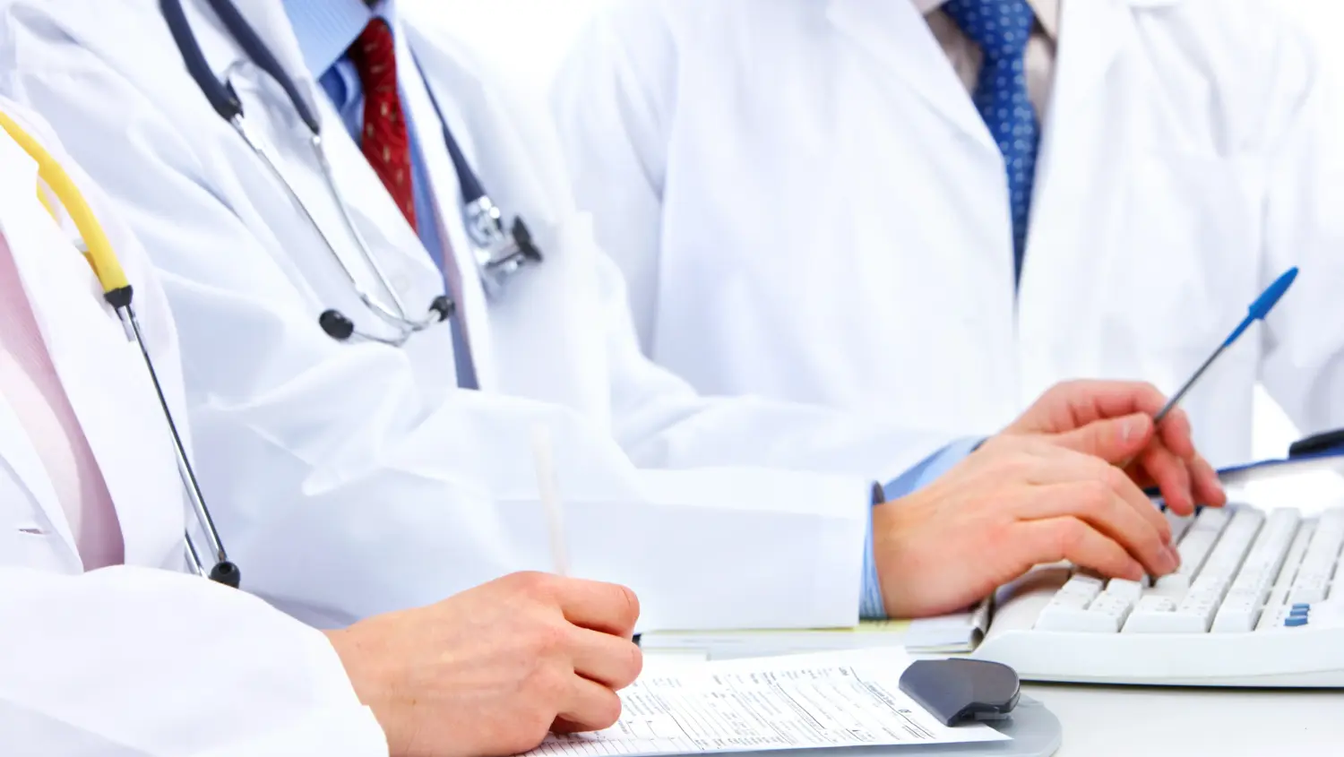 Three doctors in white coats work together at a desk, one typing on a keyboard, another writing on a clipboard, and the third holding a pen. Stethoscopes and formal clothing are visible.