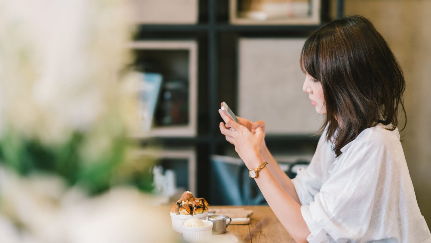 A woman with shoulder-length hair sits at a wooden table in a cafe, looking at her smartphone. In front of her are a dessert and a small cup.