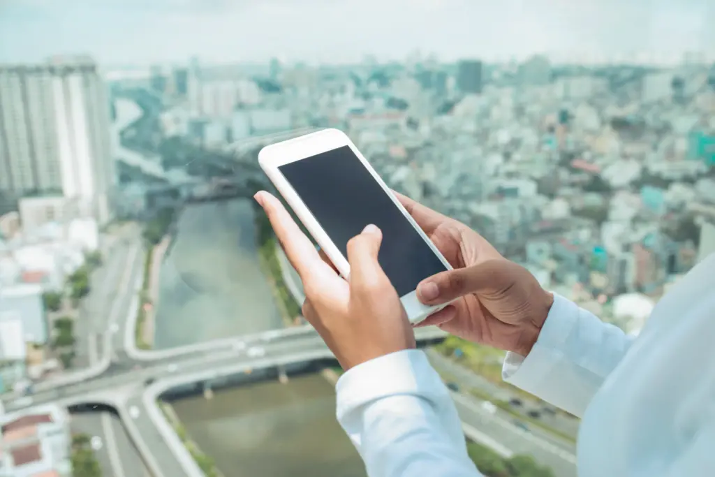 A person in a white shirt holds and uses a smartphone near a large window, with a blurred cityscape, river, and many buildings visible in the background.