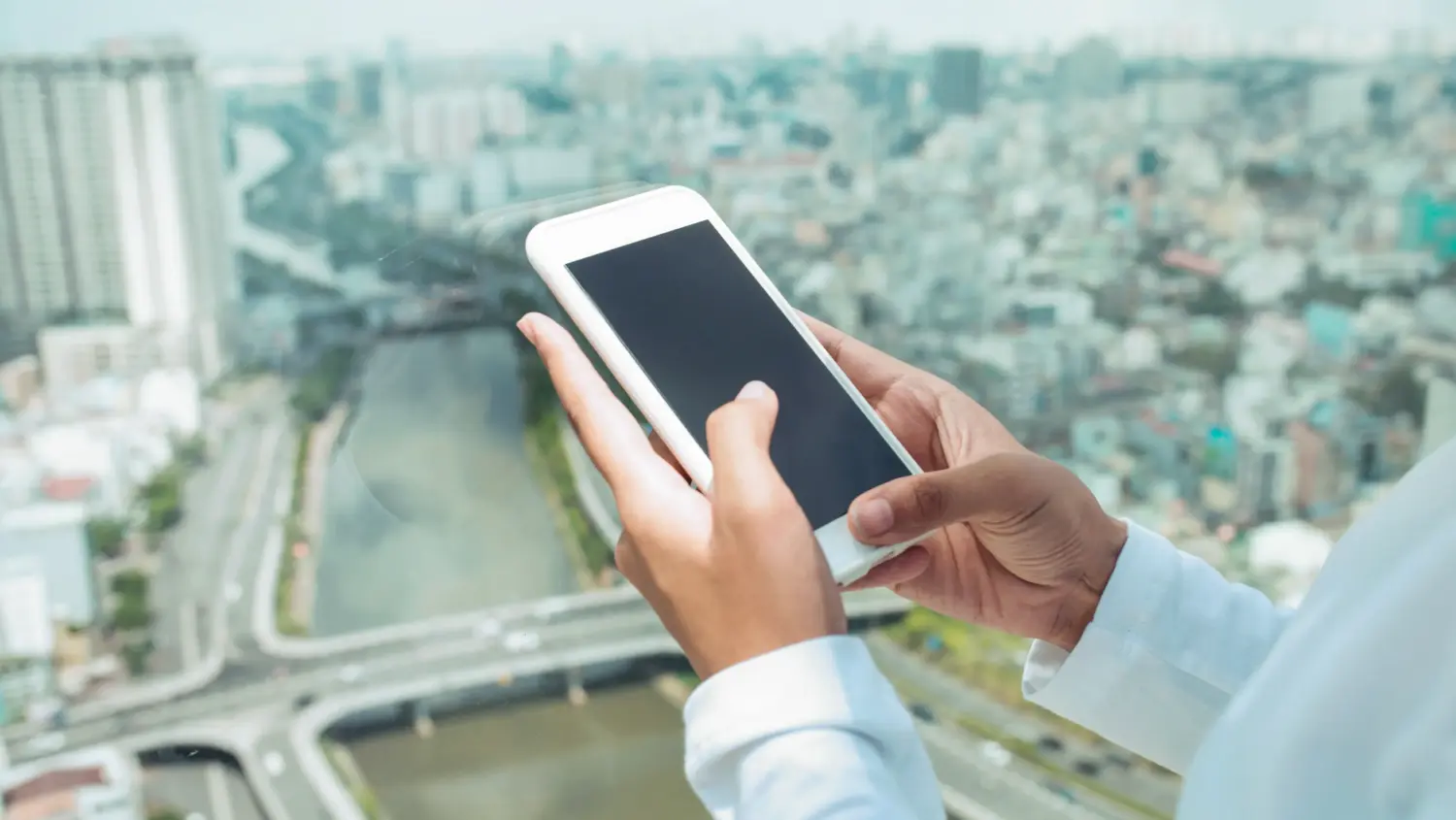 A person in a white shirt holds and uses a smartphone near a large window, with a blurred cityscape, river, and many buildings visible in the background.