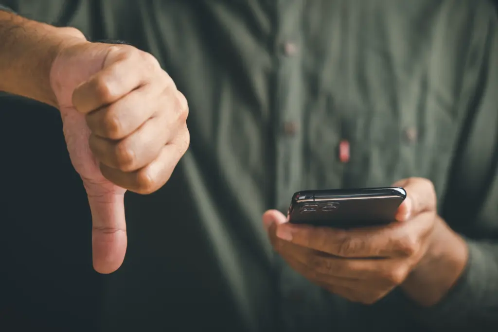 A person holding a smartphone in one hand and giving a thumbs-down gesture with the other hand, wearing a dark green shirt.