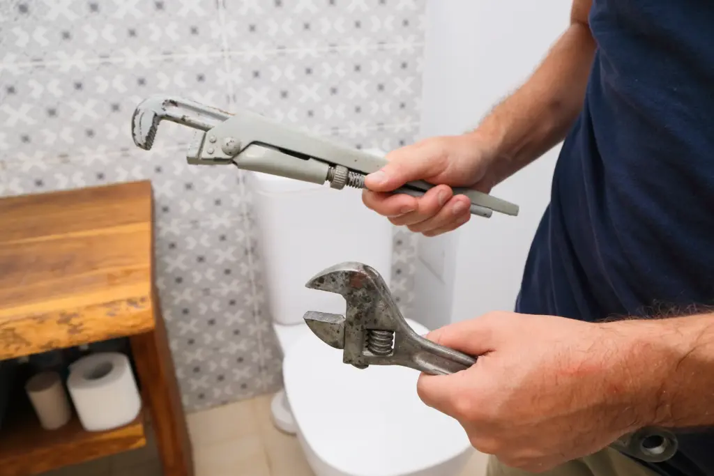 A person standing in a bathroom holds a pipe wrench in one hand and an adjustable wrench in the other, with a toilet and wooden cabinet visible in the background.