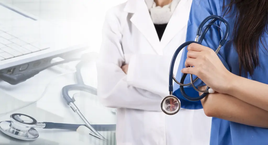 Two healthcare professionals stand together, one in a white coat with arms crossed and the other in scrubs holding a stethoscope, with medical tools and a laptop in the background.