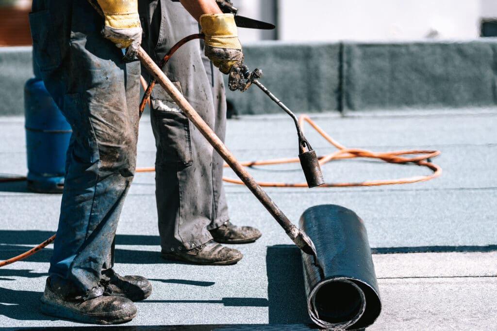 Two workers use a propane torch to apply roofing felt to a flat roof. One unrolls the felt while the other uses the flame to adhere the material to the roof surface.