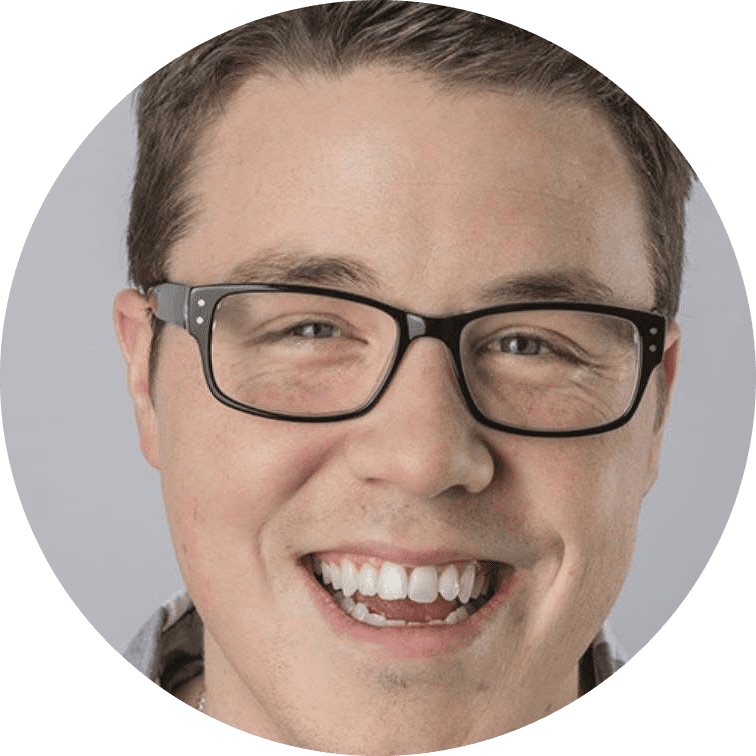 A smiling team member with short brown hair and black-rimmed glasses, showing teeth. The background is plain and light-colored.