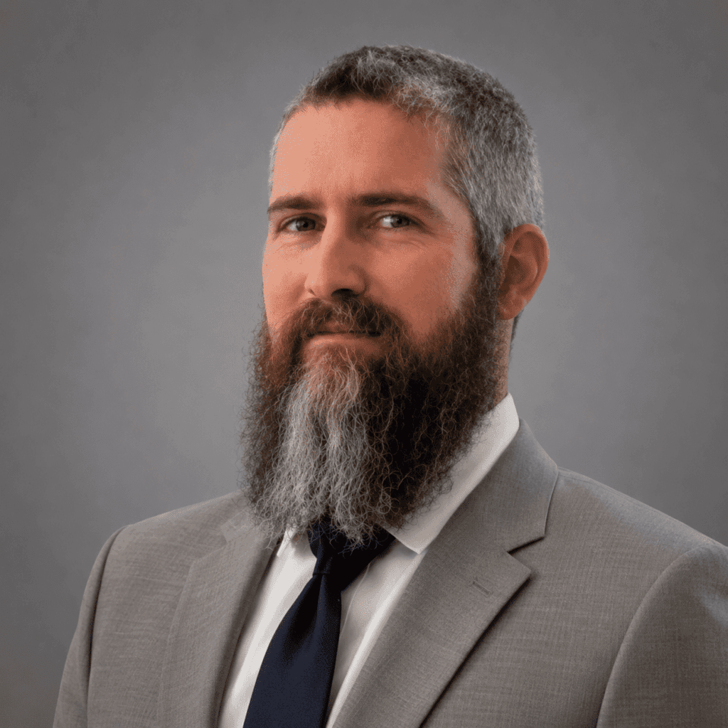 A man with a full, graying beard and short hair wearing a gray suit, white shirt, and dark tie poses against a plain gray background.
